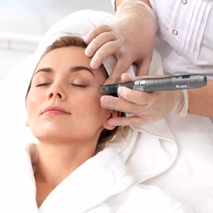 Close-up photo of a woman with a towel wrapped around her head and wearing a white robe, receiving microneedling with a skin